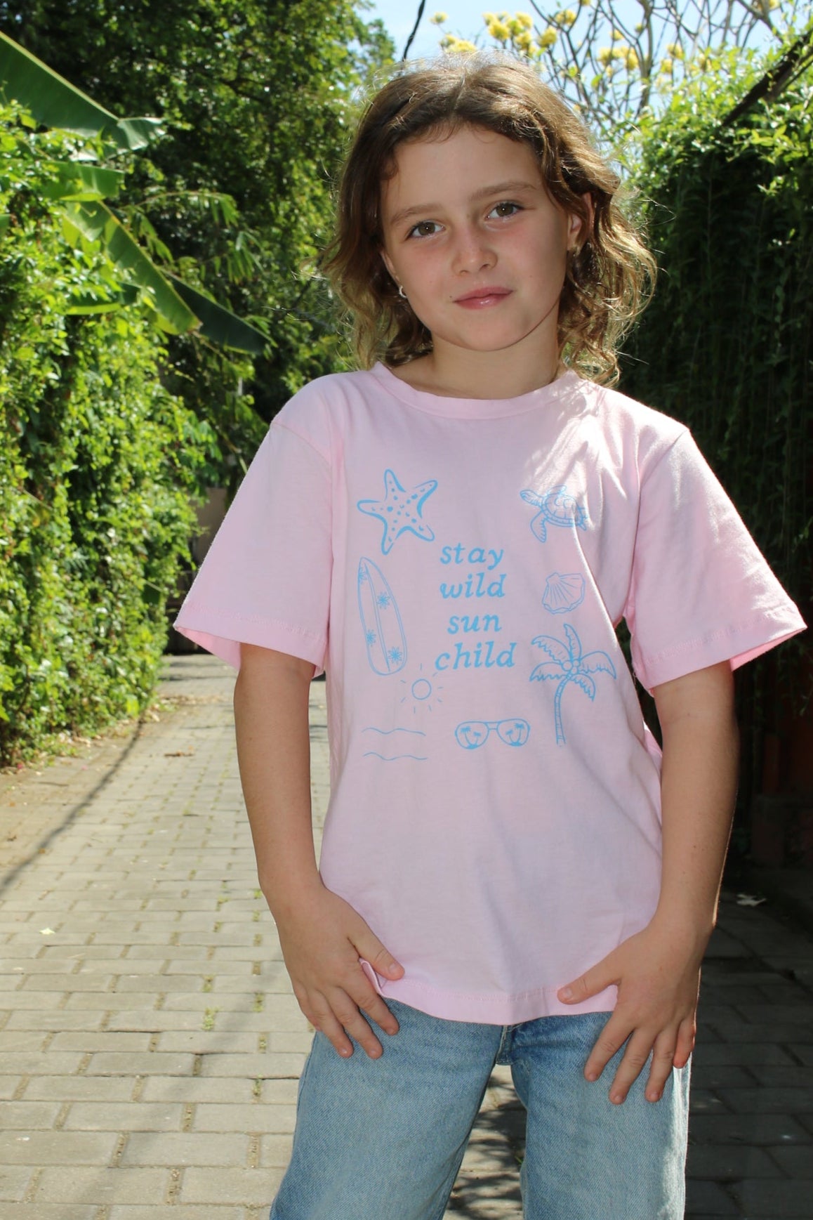 Person wearing a pink shirt with text and blue jeans standing outdoors on a sunny day.