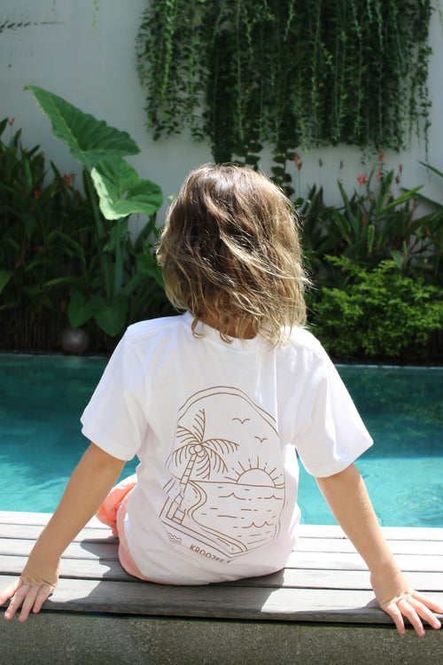child wearing a white shirt with a graphic design, standing in front of green plants by a pool.