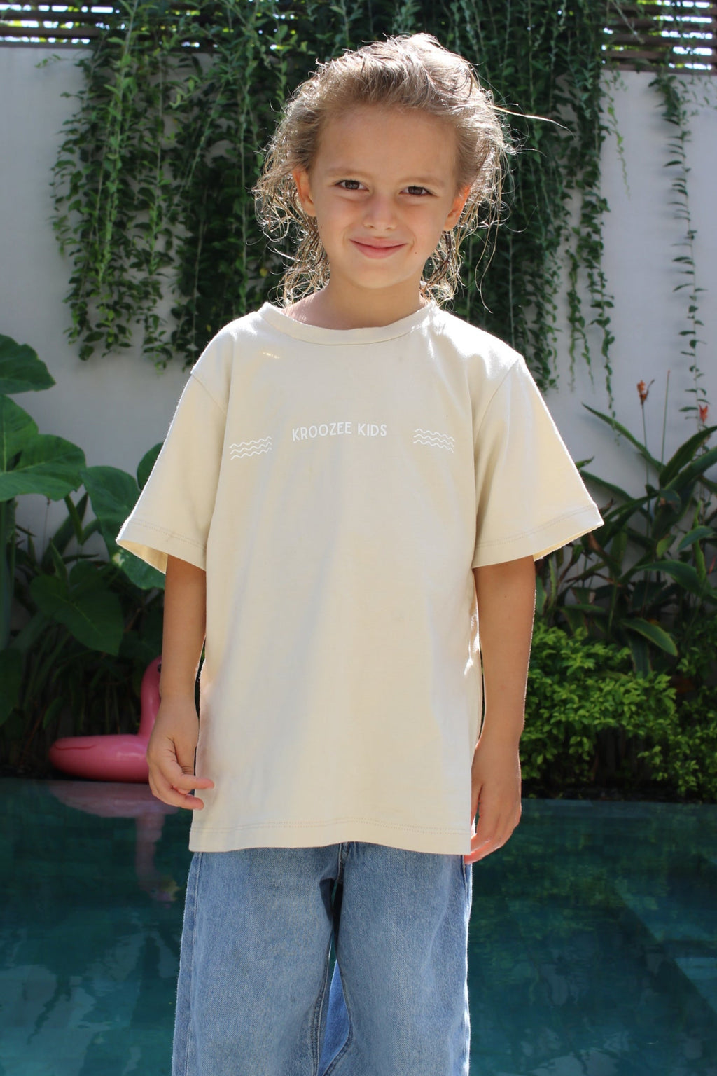 Person wearing a light-colored shirt and jeans standing outdoors with plants in the background