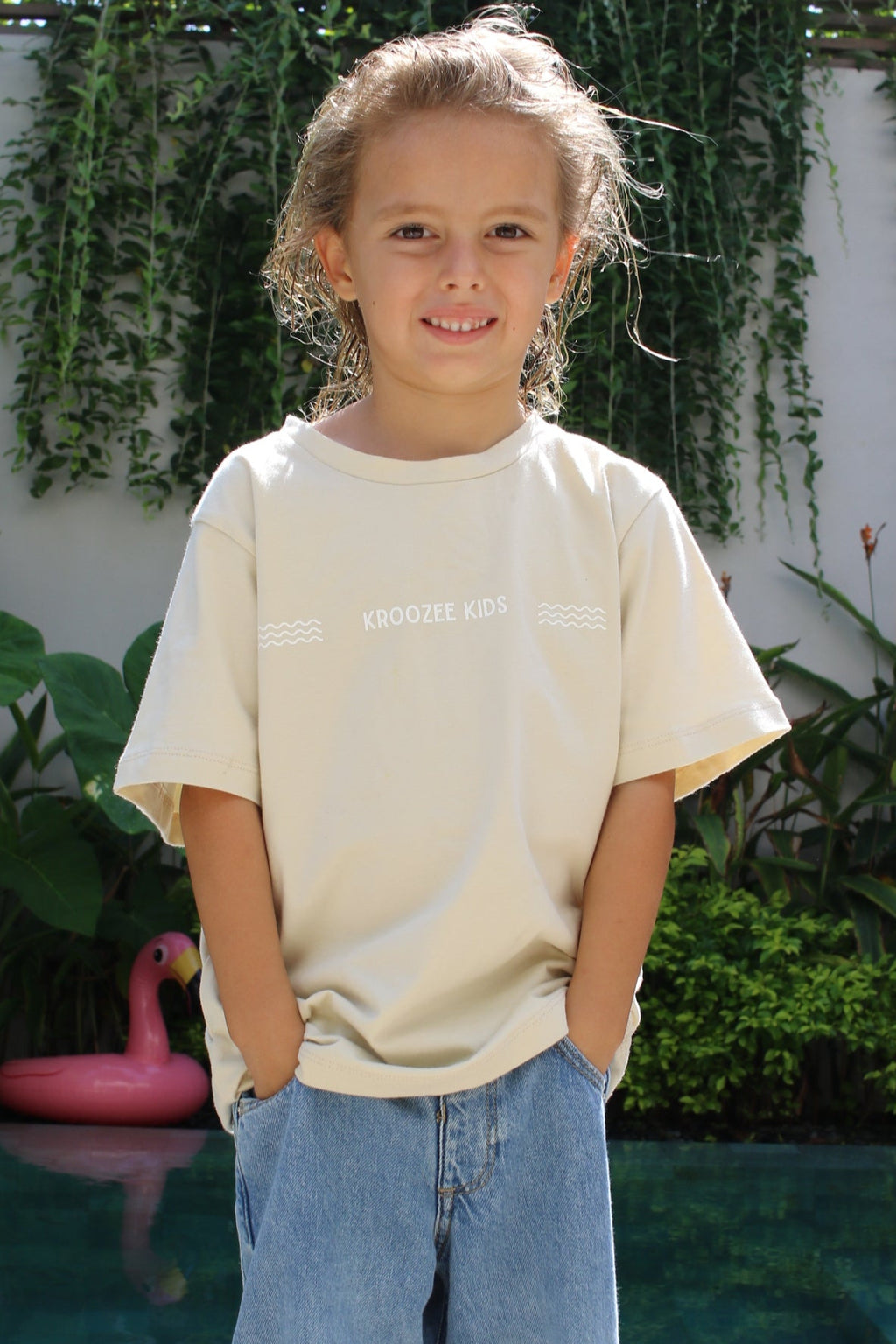 Person wearing a white top and blue jeans standing outdoors with greenery in the background