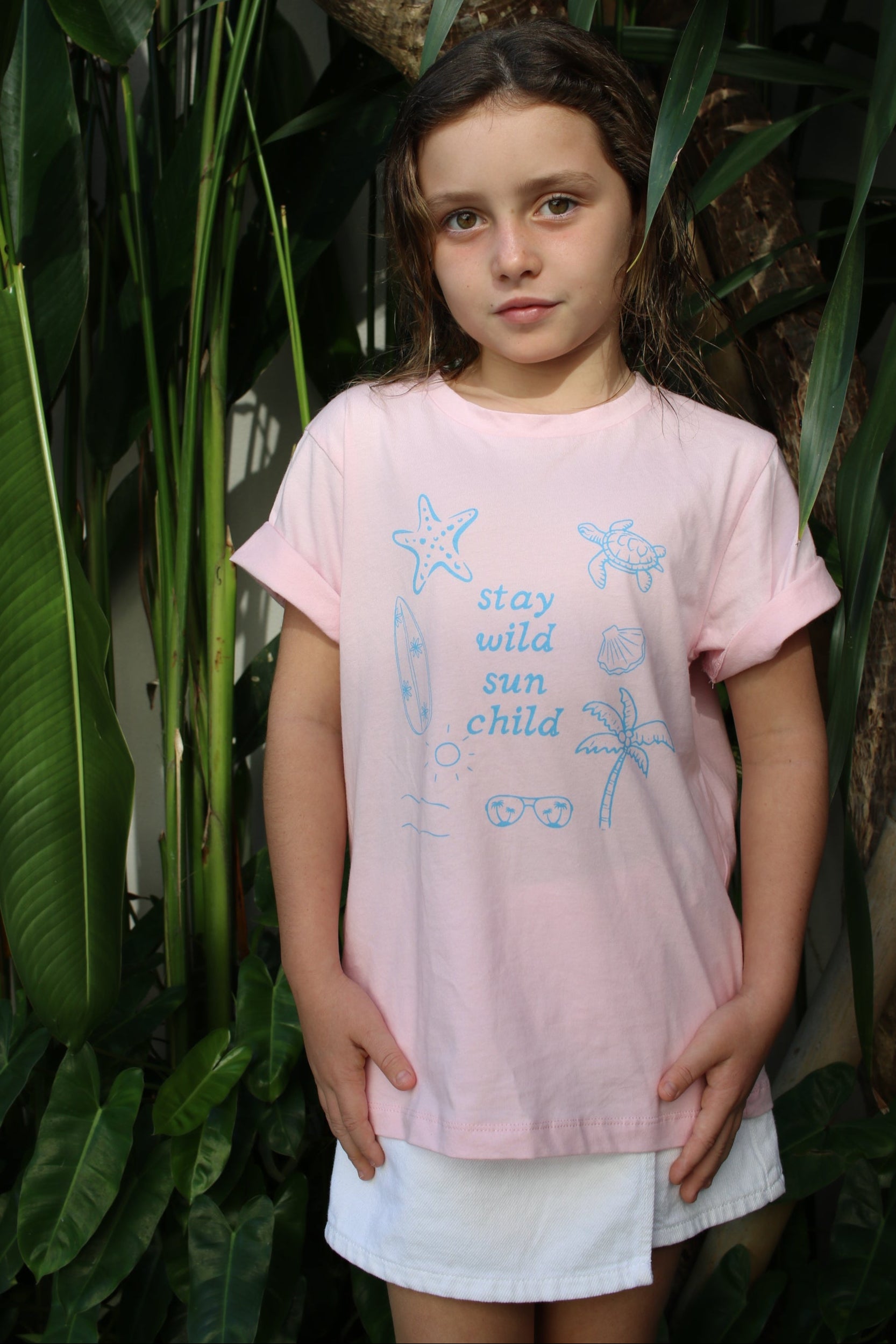 Child wearing a pink shirt with blue designs and text, standing among green plants.