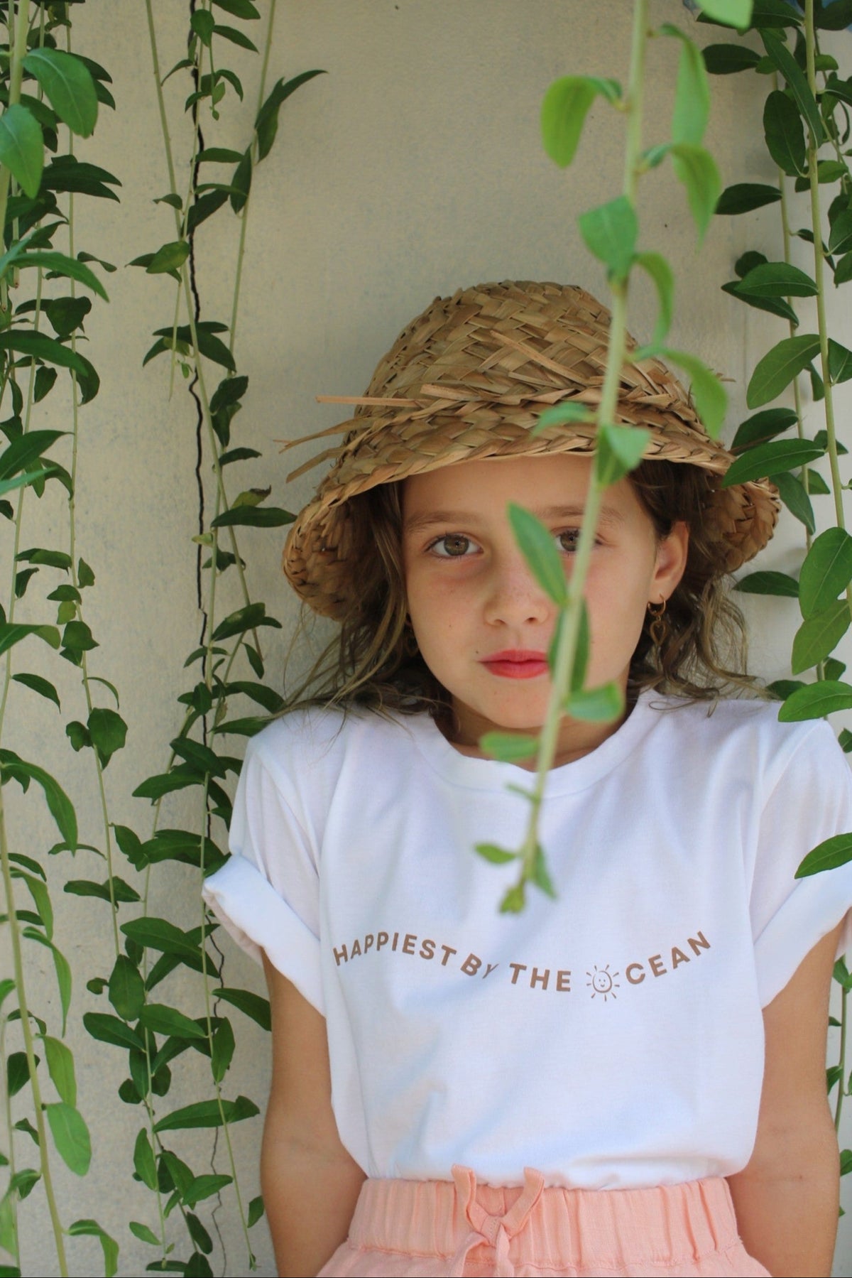 Young girl wearing a straw hat and white t-shirt with text, standing among green plants.